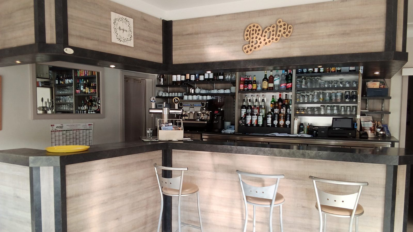 Le comptoir du bar en angle, ses trois tabourets hauts, la machine à café et la tireuse ; derrière, les étagères de bouteilles et de verres sous l’enseigne « Bar » en lettres de bois.