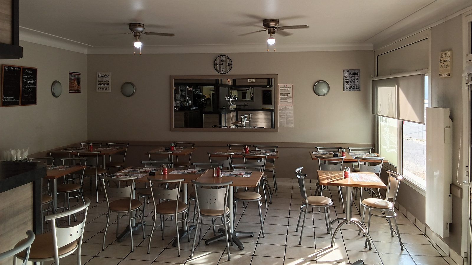 La salle de restaurant et ses tables carrées dressées de sets de table, sous deux ventilateurs de plafond ; au fond, l’ardoise des desserts et le passe de la cuisine, et à droite la fenêtre sur la rue.