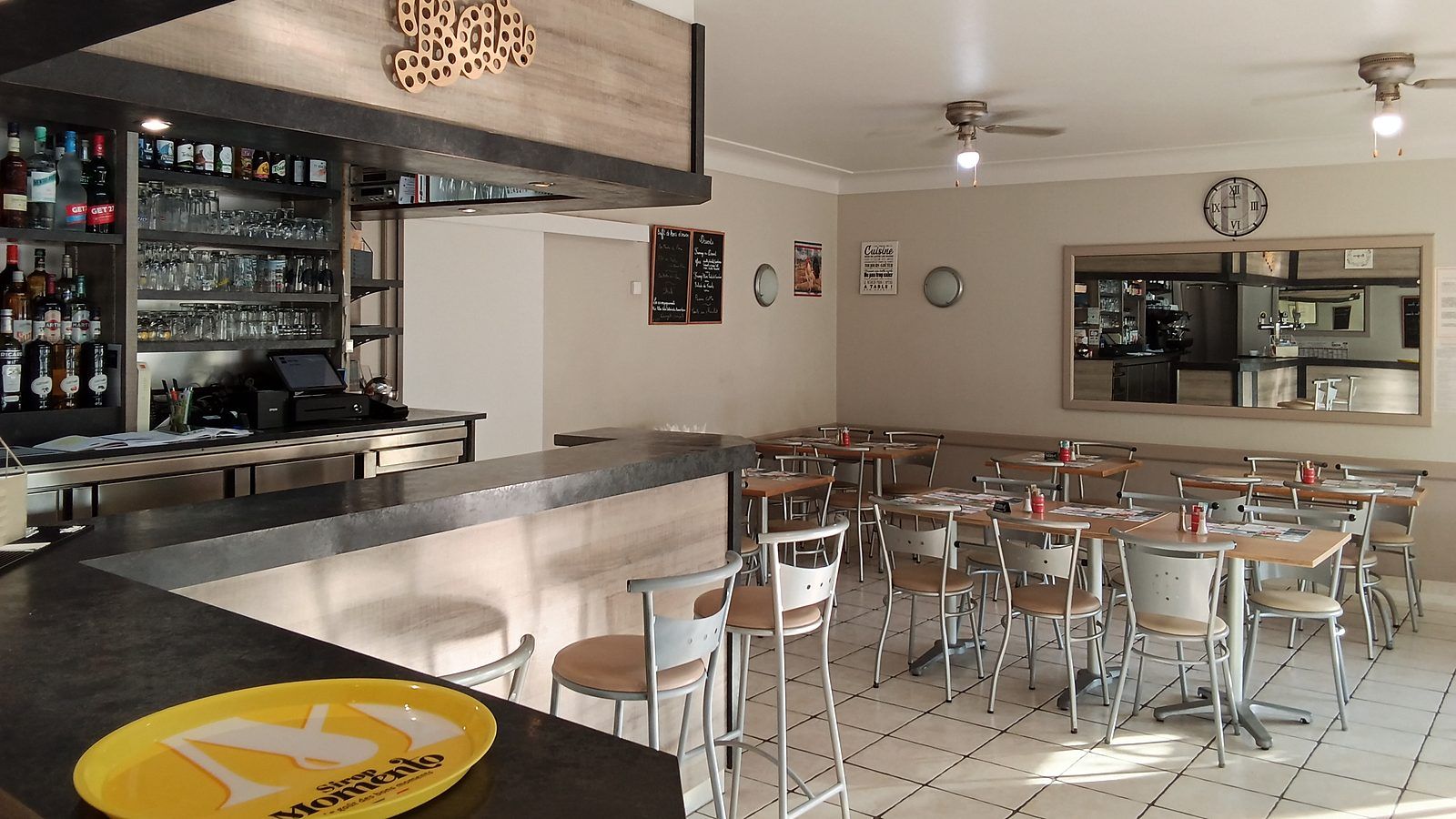 La salle vue depuis le comptoir : le bar et ses bouteilles au premier plan à gauche, les tables et leurs chaises qui s’étendent vers le fond, et l’horloge murale au-dessus.